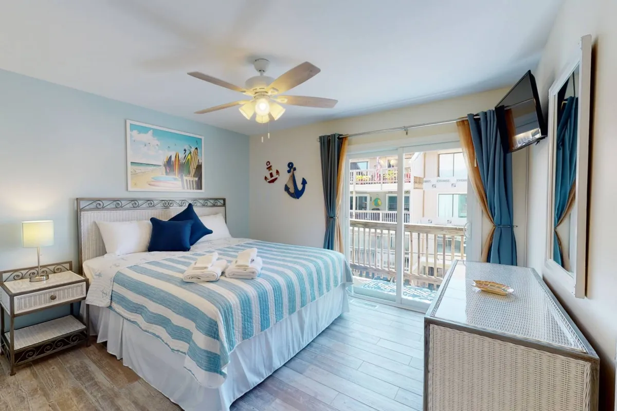 A cozy bedroom with blue and white bedding, beach-themed wall art, and nautical accents. A large window offers balcony access, filling the room with natural light and providing a sense of openness and relaxation.