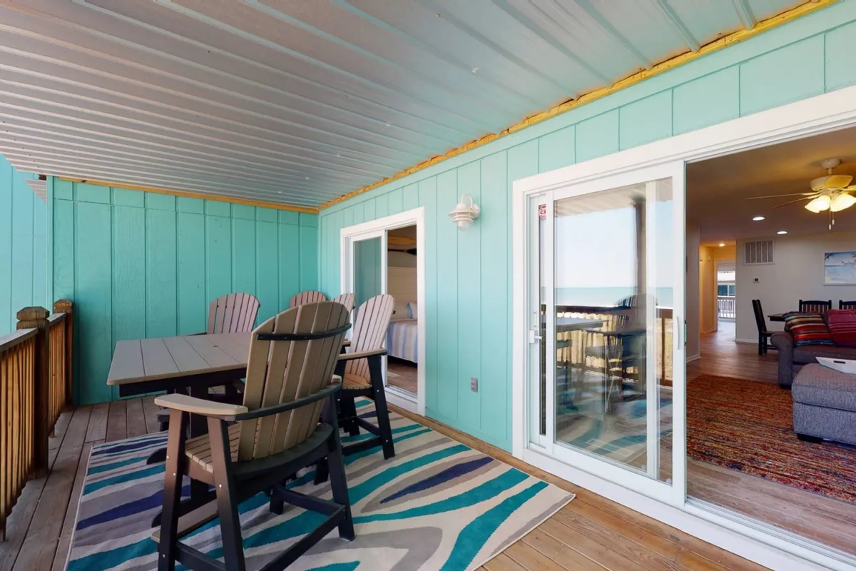 An inviting outdoor dining space featuring a large dining table with ample seating and adirondack chairs. The area is set against blue walls, with a rug featuring a wavy design, and sliding glass doors connecting to the interior. The view suggests a relaxed atmosphere ideal for group meals and socializing, and there is an ocean or deck view visible, reinforcing the alfresco dining experience.