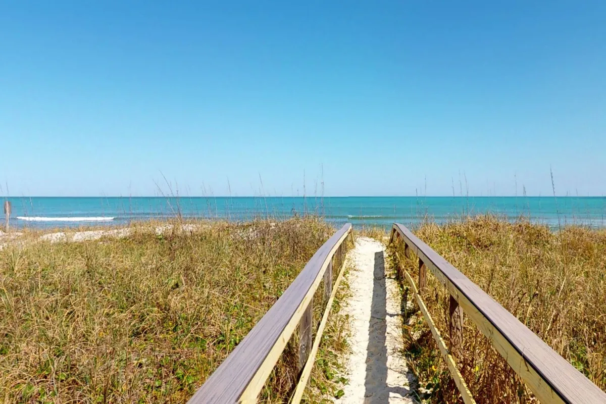 A serene wooden walkway bordered by dune grass leads directly out to the ocean, with wide sandy beaches visible beyond. The tranquil mood and open view evoke a sense of direct access and immediate immersion in the beach environment.