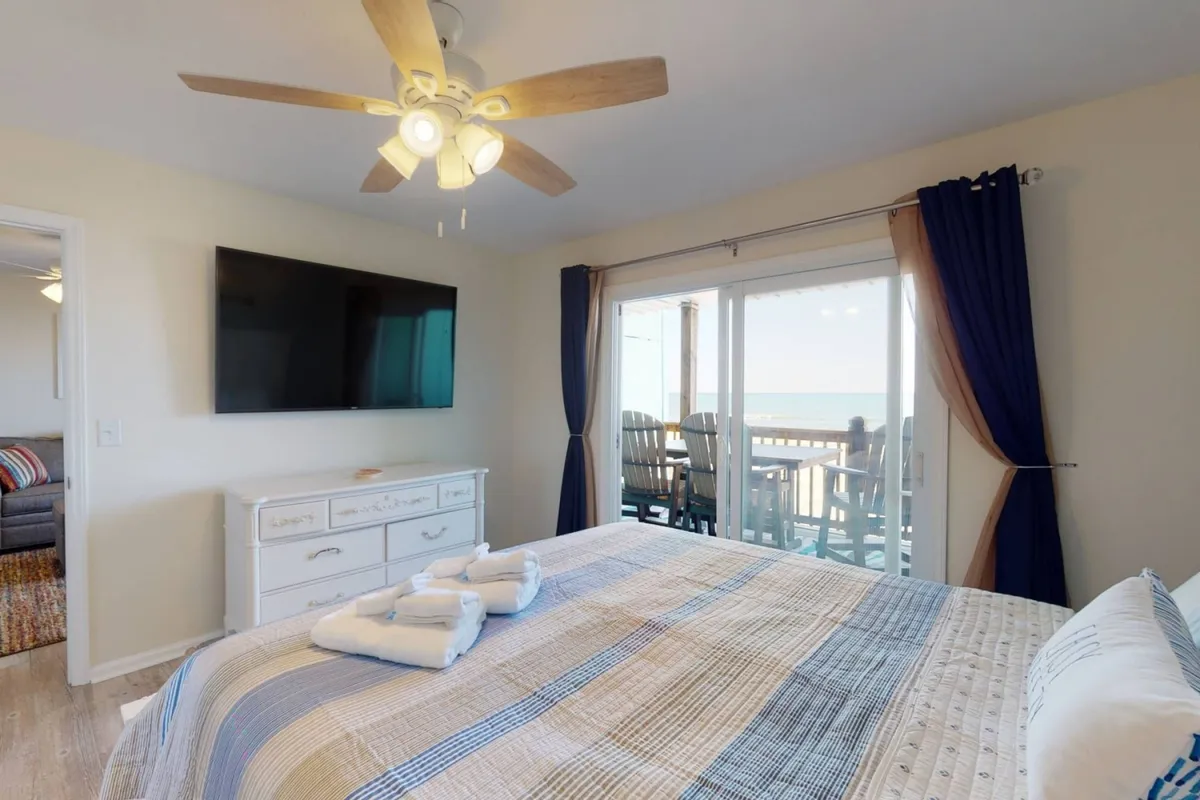 A serene bedroom with a large bed featuring a patterned blanket faces floor-to-ceiling windows that offer sweeping ocean views. A large TV is mounted on the wall, and comfortable seating on the patio is visible through the glass, bathed in natural light.