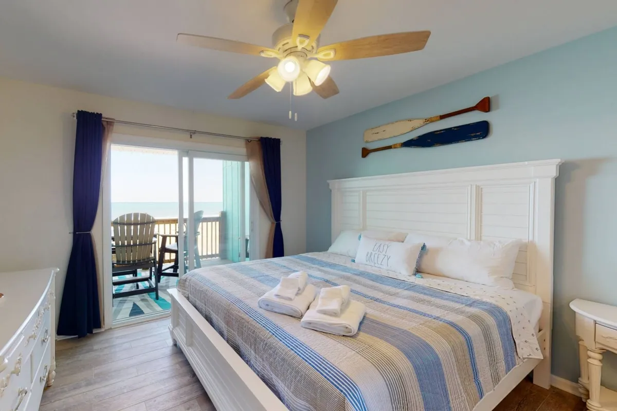 A king-sized bed faces decorative wall paddles, flanked by elegant drapes and a balcony view. The room is softly lit, inviting relaxation and providing a sense of privacy and retreat.