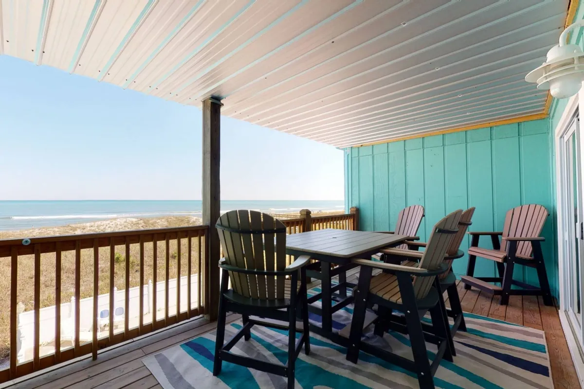 A tranquil scene showcasing a dining table and adirondack chairs set on a deck overlooking the ocean. The setup faces out toward a serene ocean view, suggesting a perfect spot for morning coffee, meals, or sunset watching. The mood is peaceful, with a focus on enjoying the coastal backdrop while dining outdoors.