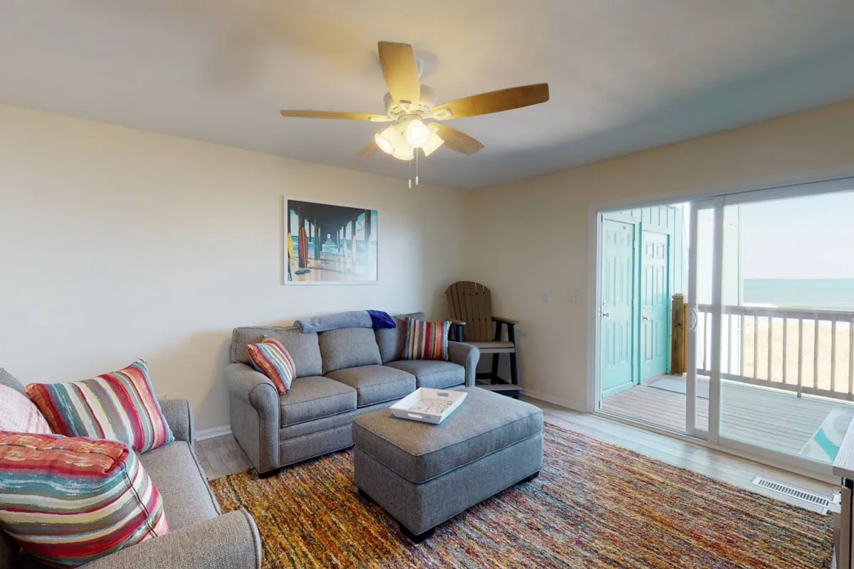 A spacious living area with a comfortable sofa and ottoman, bright artwork, and a door leading to a balcony. The room feels inviting and comfortable, perfect for gathering or accommodating extra guests with a pullout bed.