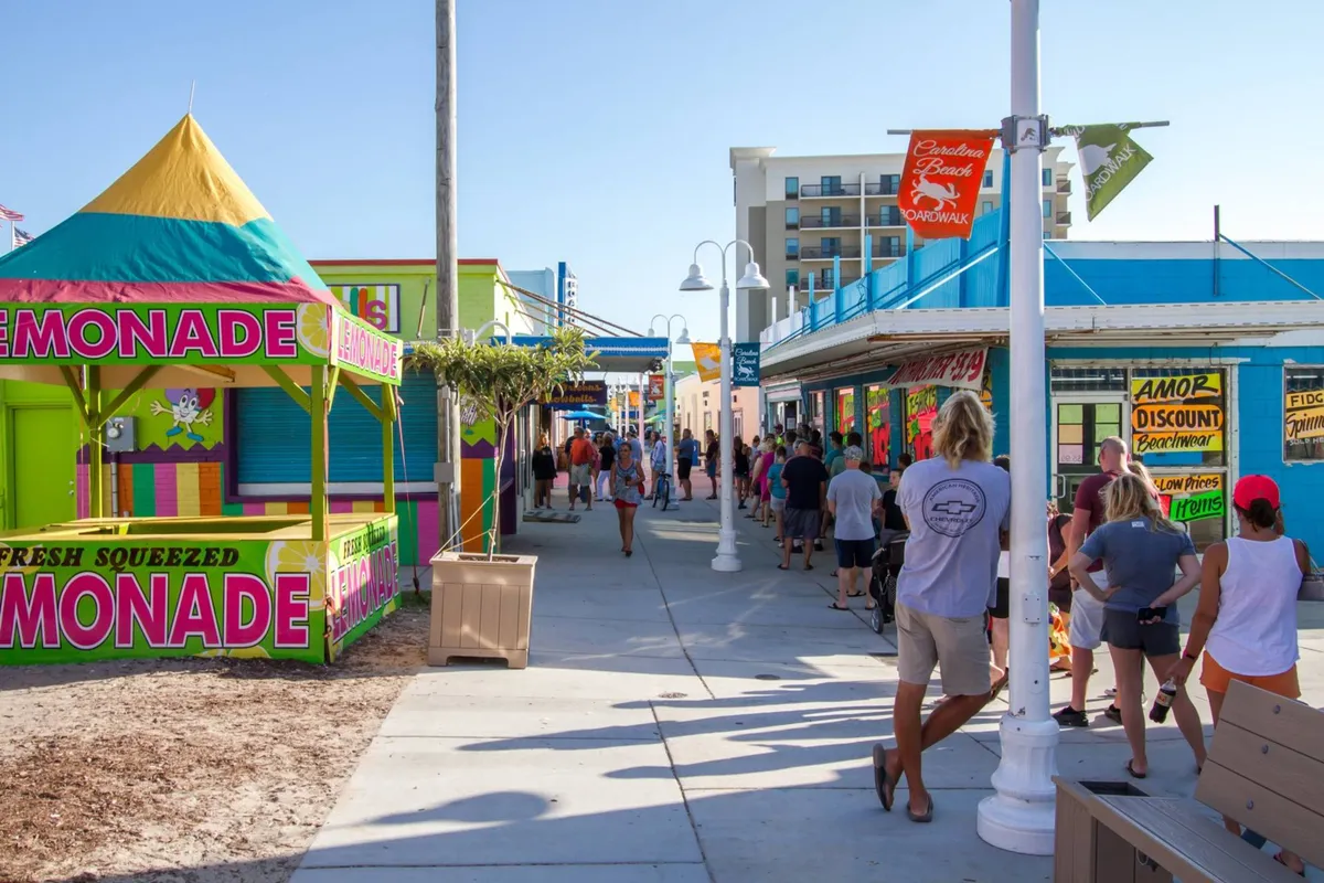 A lively boardwalk bustling with families and children, featuring a colorful lemonade stand, various shops, and a cheerful crowd. The scene is full of energy and excitement, capturing the essence of local entertainment and activities.