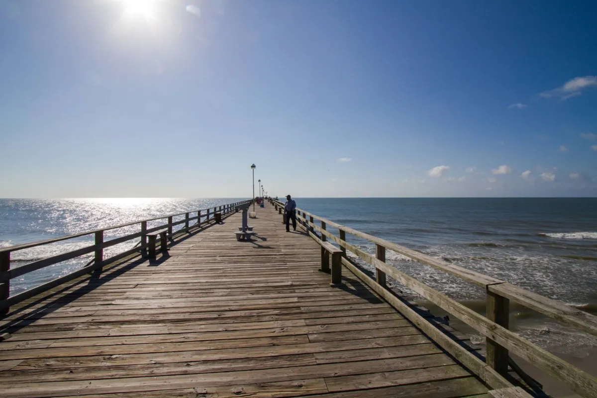A serene shot of a wooden pier stretching over the ocean under a glowing sky, with distant figures basking in the sunset ambiance. The scene evokes tranquility, intimacy, and the joy of sharing quiet moments by the water.