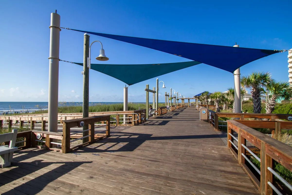 A vibrant wooden boardwalk lined with colorful shade sails and palm trees, overlooking the sparkling ocean. People are seen walking and enjoying the relaxed, beachside environment.