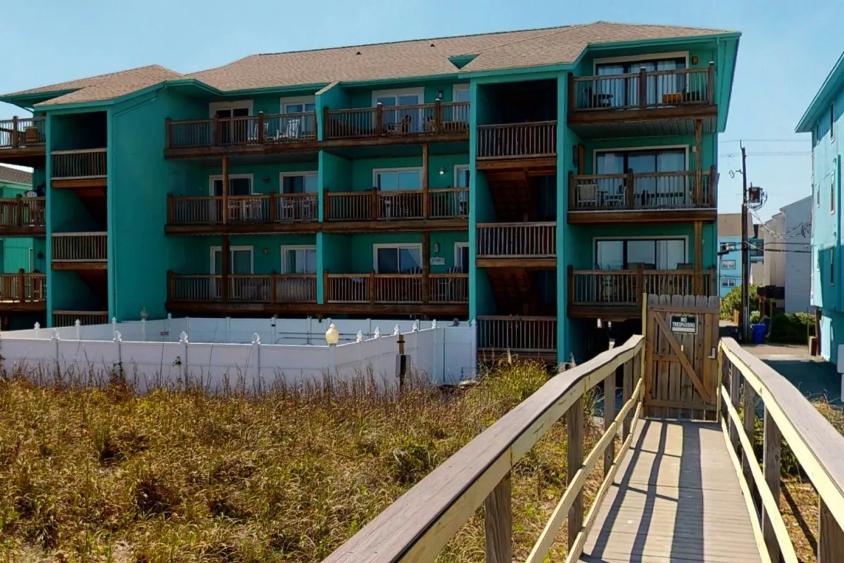 The Easy Breezy property’s teal exterior with wooden balconies overlooks a relaxing pathway leading towards the beach, capturing the connection between private decks and the ocean beyond. The balconies suggest spaces for quiet coffee moments or sunset viewing for couples.