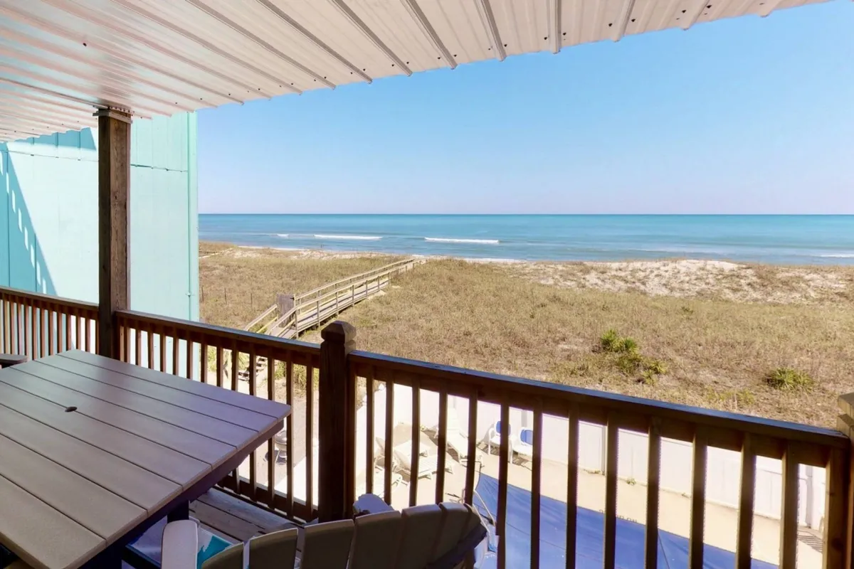 The hero image captures a breathtaking ocean view from a private wooden deck overlooking Carolina Beach. Rolling sand dunes lead the eye to the blue horizon, while comfortable deck seating invites guests to relax and soak up the peaceful coastal ambiance.