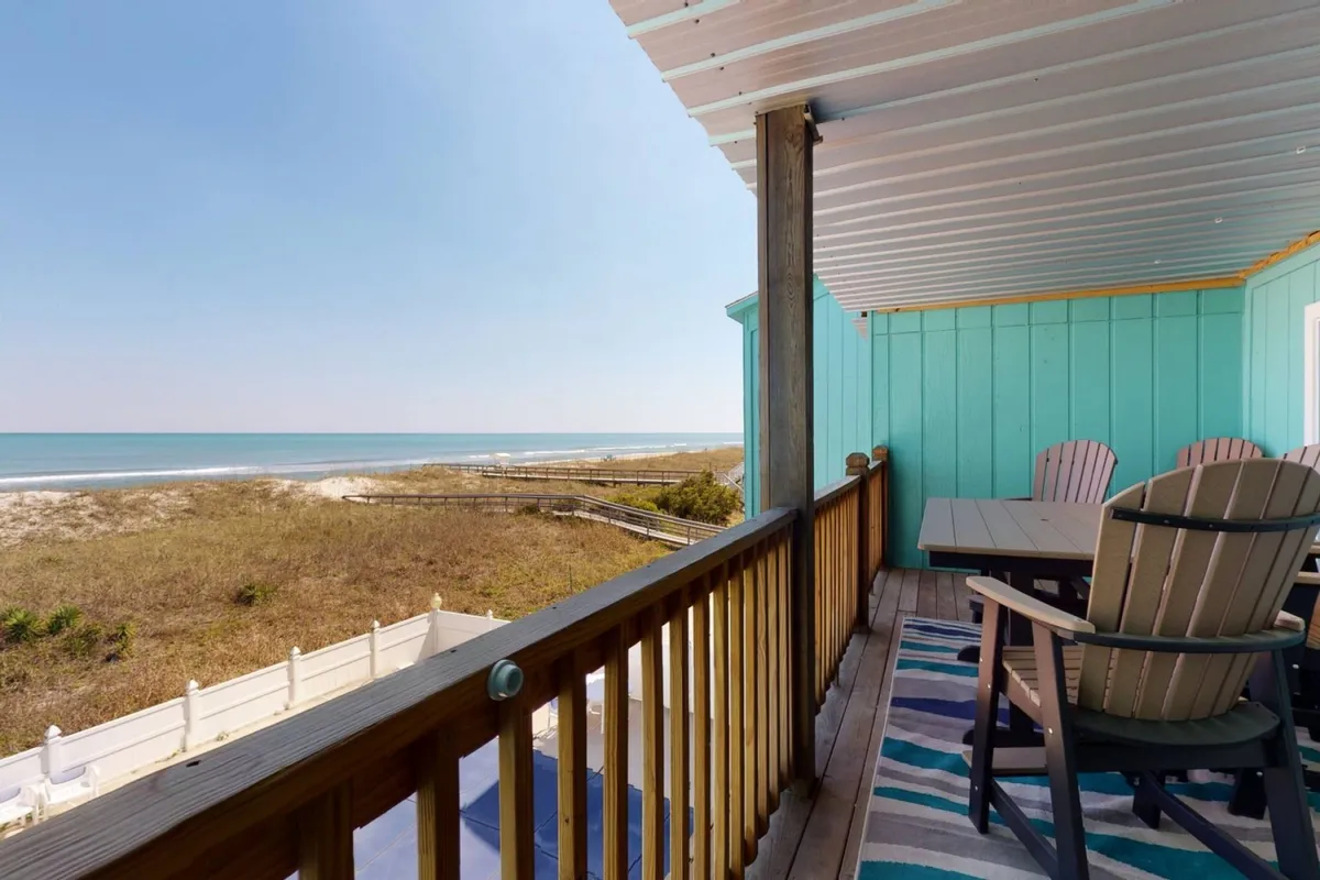 A private deck with outdoor seating, a wooden railing, and a striped rug, all overlooking expansive ocean views. The scene is bathed in natural light, inviting relaxation, dining, or reading with the soothing sound of waves in the background.