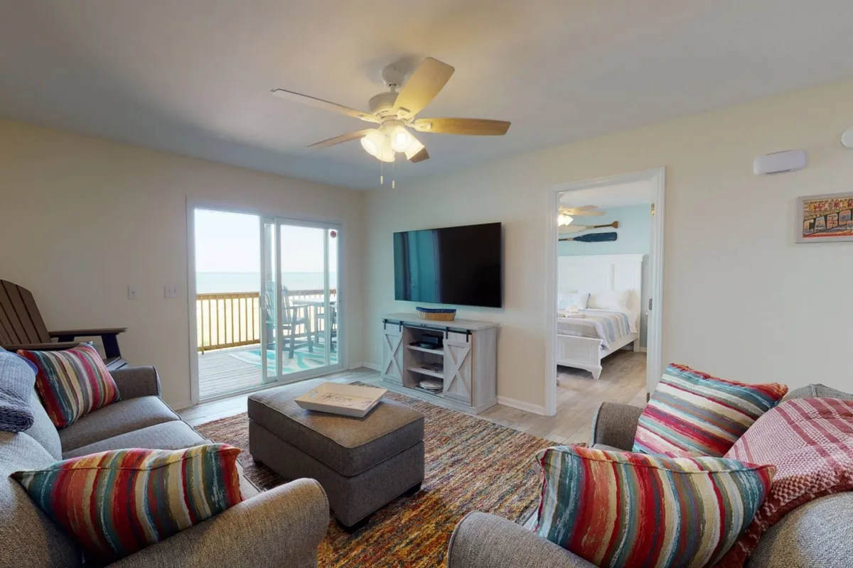A welcoming living room with a plush sofa adorned with colorful cushions, a stylish coffee table, a large television for entertainment, and a sliding glass door that opens to a deck. Natural light pours in, highlighting the open layout and providing a glimpse of the outdoor space and possible ocean views beyond.